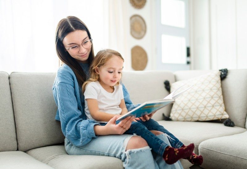 What To Know Before Babysitting a Younger Relative 1 A teen babysitting her younger relative. The little girl is sitting on her lap, reading a book that the teen is holding open.