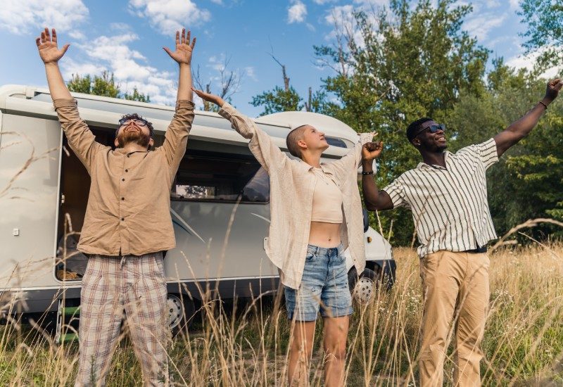 Is Van Life Really As Freeing as It Looks? 1 A trio of friends standing outside of their white van, holding their arms out to the air as they stretch and smile.